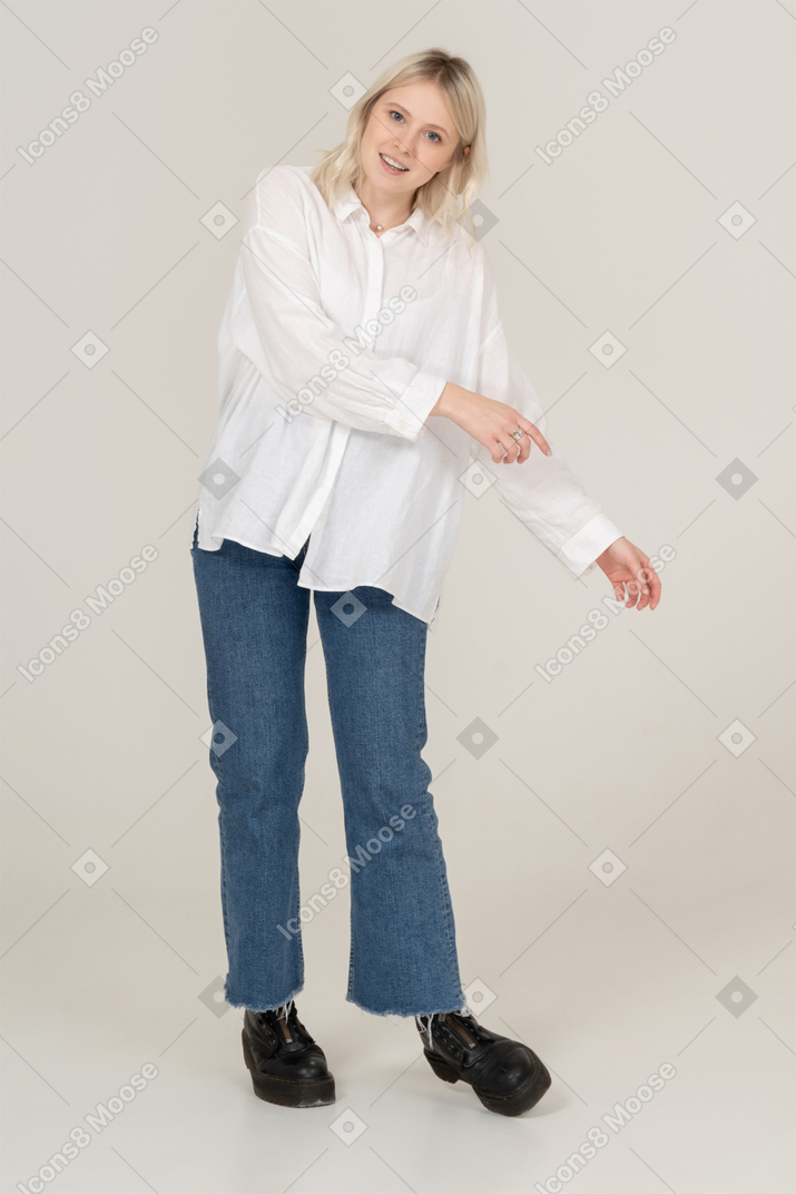 Front view of a blonde female in casual clothes stepping aside while outstretching hands and looking at camera