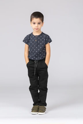 Front view of a cute boy in casual clothes posing with hands in pockets and looking at camera