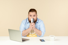 Young overweight man sitting at the table and holding bank card close to his mouth