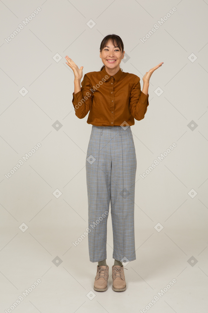 Vista frontal de uma jovem mulher asiática encantada de calça e blusa, levantando as mãos