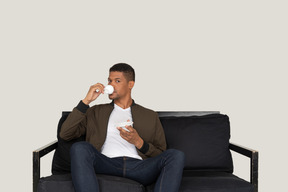 Front view of a young dreaming man sitting on a sofa while drinking coffee