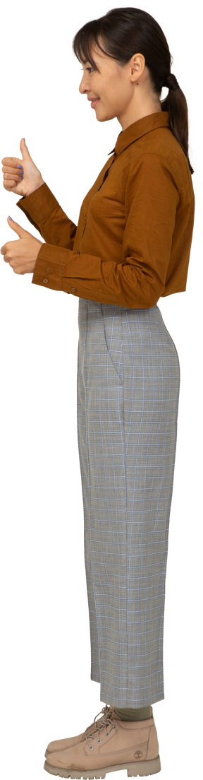 Vista lateral de uma jovem mulher asiática de calça e blusa mostrando os polegares para cima