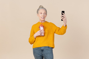 Old woman holding plastic cup to go and making selfie