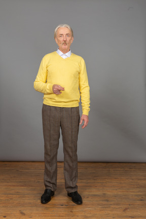 Vista frontale di un vecchio perplesso in un pullover giallo alzando la mano e guardando la fotocamera
