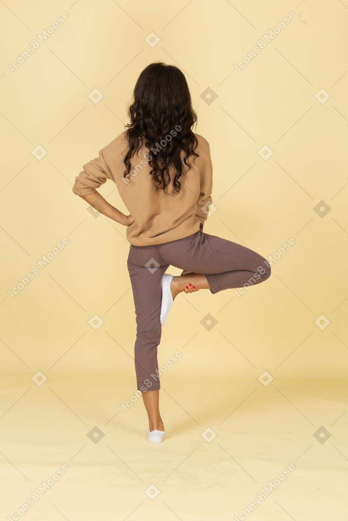 Vista posterior de una mujer joven de piel oscura poniendo la mano en la cadera mientras levanta la pierna