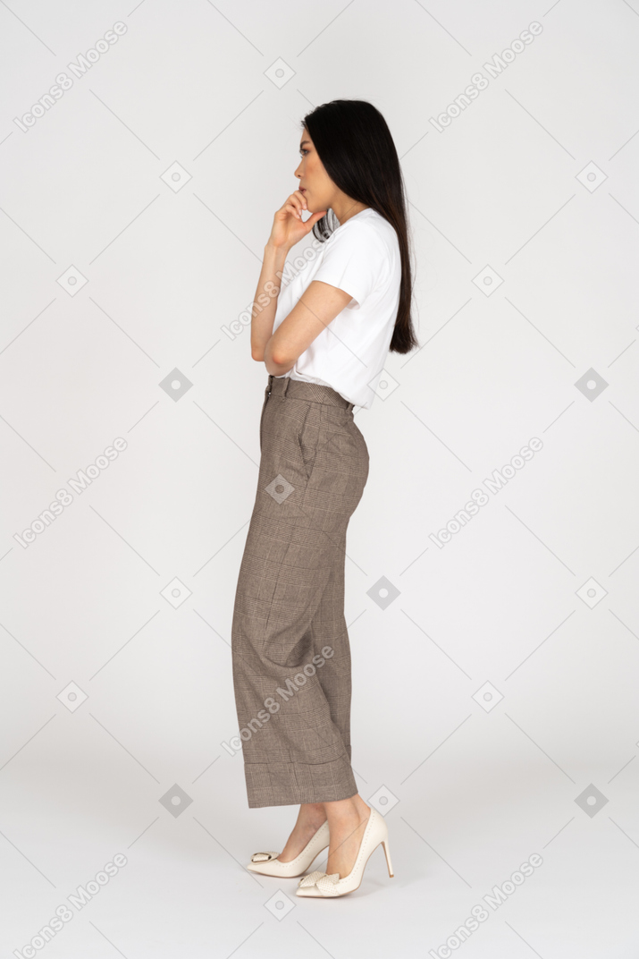Vista lateral de uma jovem atenciosa de calça e camiseta tocando o queixo