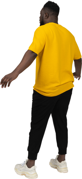 노란 티셔츠를 입고 팔을 벌리고 있는 충격을 받은 검은 피부의 젊은 남자의 4분의 3 뒷모습