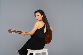 Vista traseira de uma jovem sentada de terno preto segurando o violão enquanto se vira