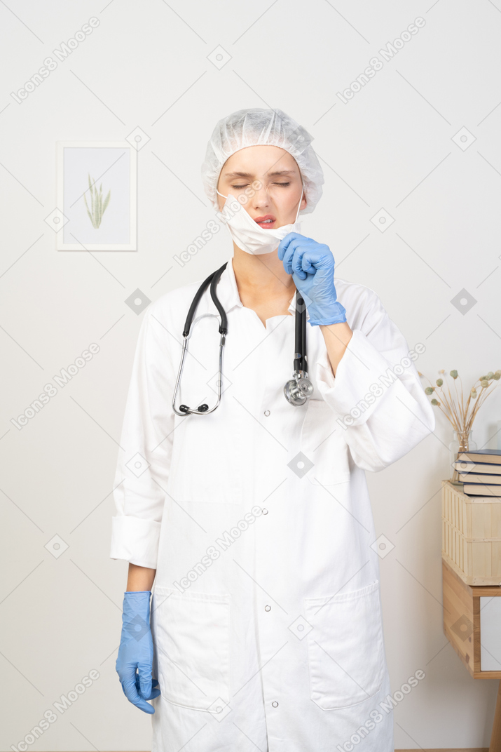 Front view of a displeased young female doctor trying to take mask off