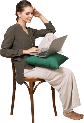 Three-quarter view of a young woman wearing home clothes sitting on a chair with a laptop and touching forehead