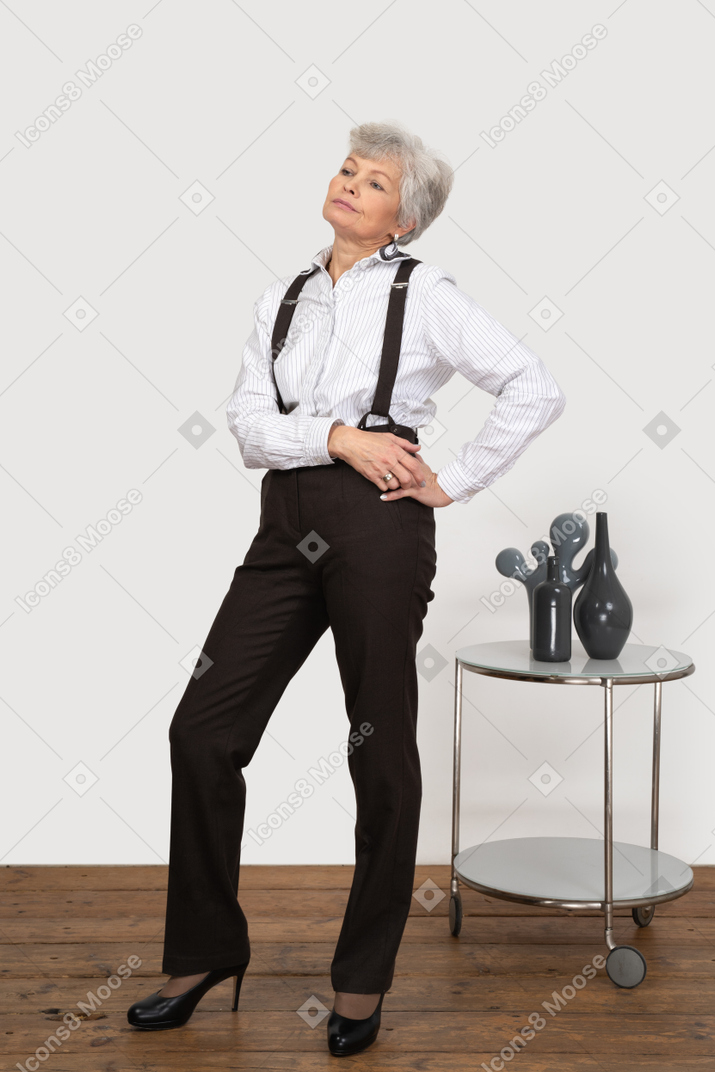 Вид в три четверти высокомерной старушки в офисной одежде, положившей руки на бедро