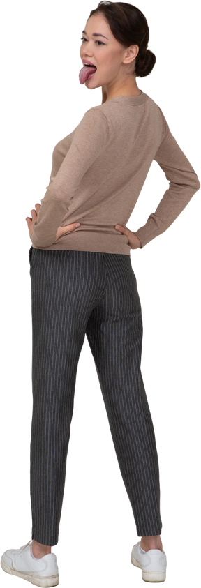 Vista posterior de tres cuartos de una joven en suéter y pantalones poniendo las manos en las caderas y mostrando la lengua