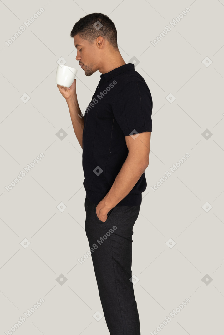 Standing in profile young man in black t-shirt and pants drinking coffee