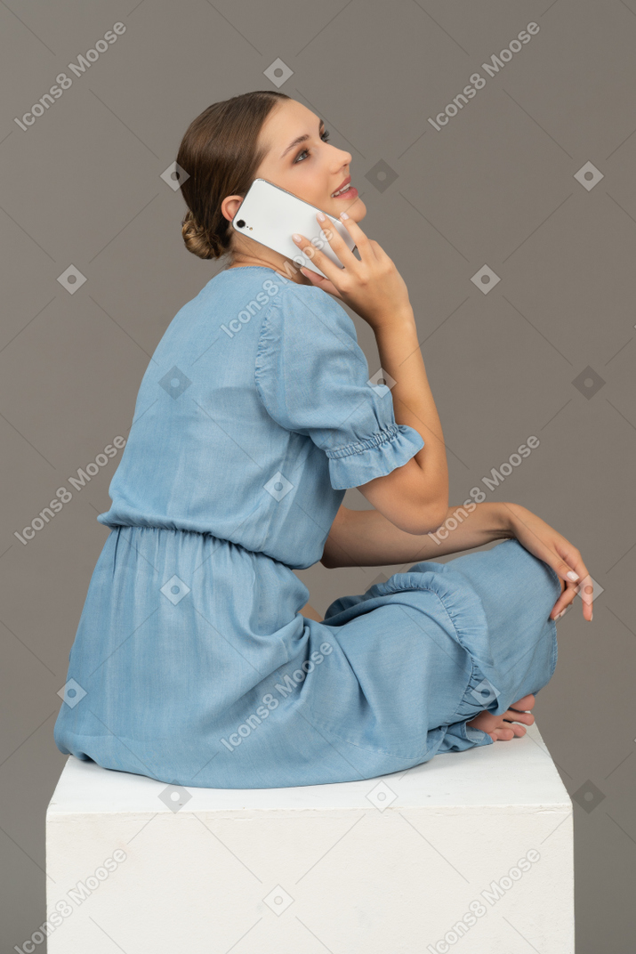 Vista lateral de uma jovem sentada no cubo e falando no smartphone