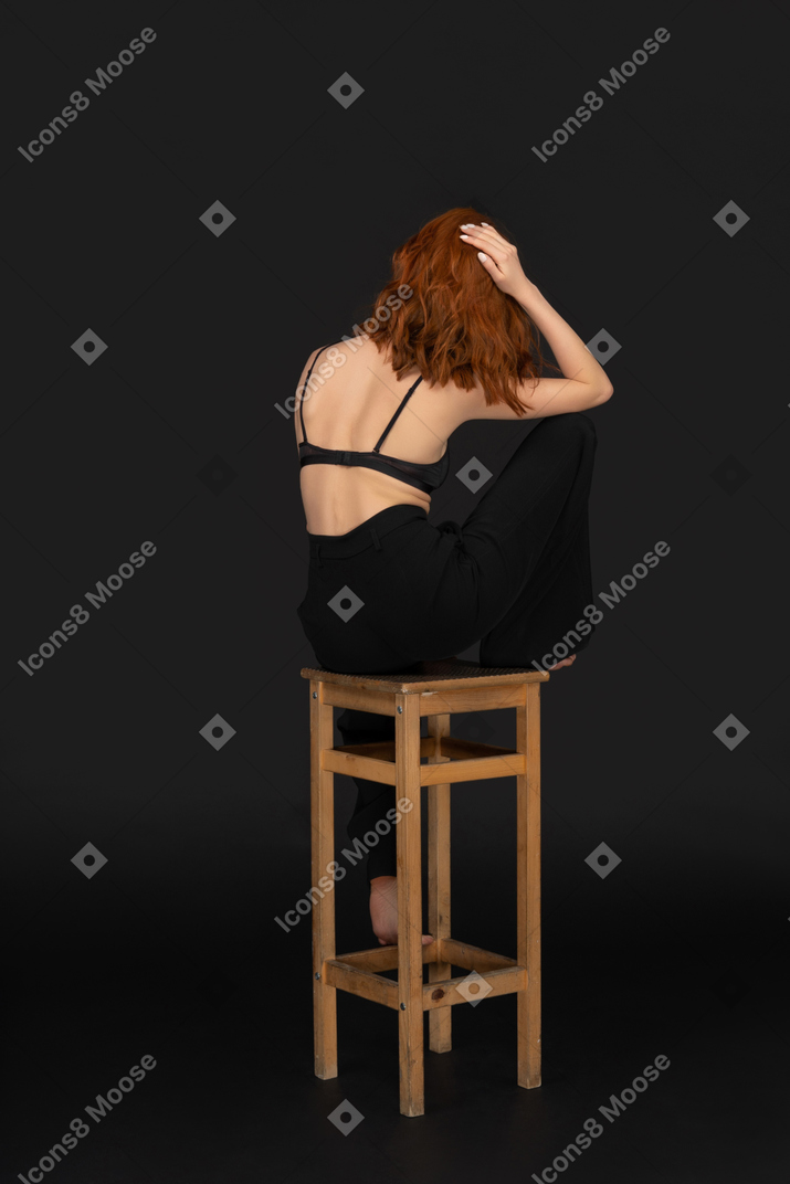 A three-quarter view of the beautiful woman dressed in black pants and bra, sitting on the wooden chair and holding her hand on the hair