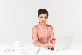Молодой индийский офисный работник работает на ноутбуке и смотрит прямо в камеру