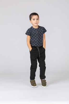Vue de face d'un garçon mignon dans des vêtements décontractés posant avec les mains dans les poches et regardant la caméra