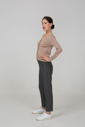 Vista di tre quarti di una giovane donna in pullover e pantaloni che mette le mani sui fianchi e mostra la lingua