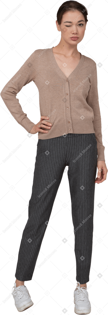 Vista frontal de una señorita guiñando un ojo en suéter y pantalones poniendo la mano en la cadera