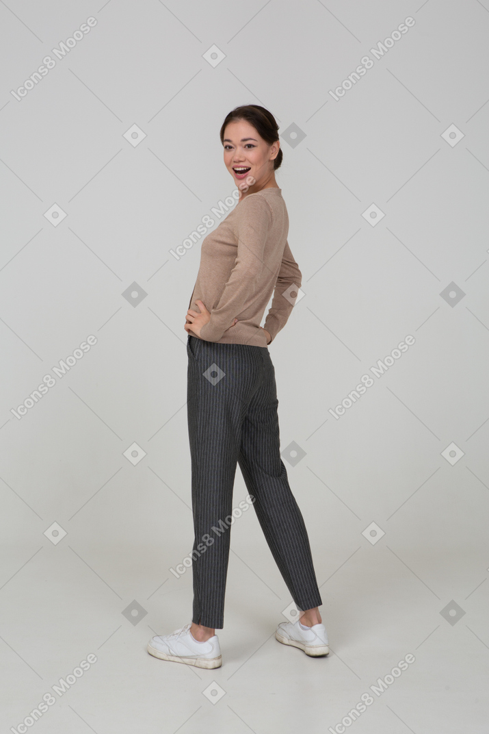 Three-quarter back view of a smiling young lady in pullover and pants putting hands on hips