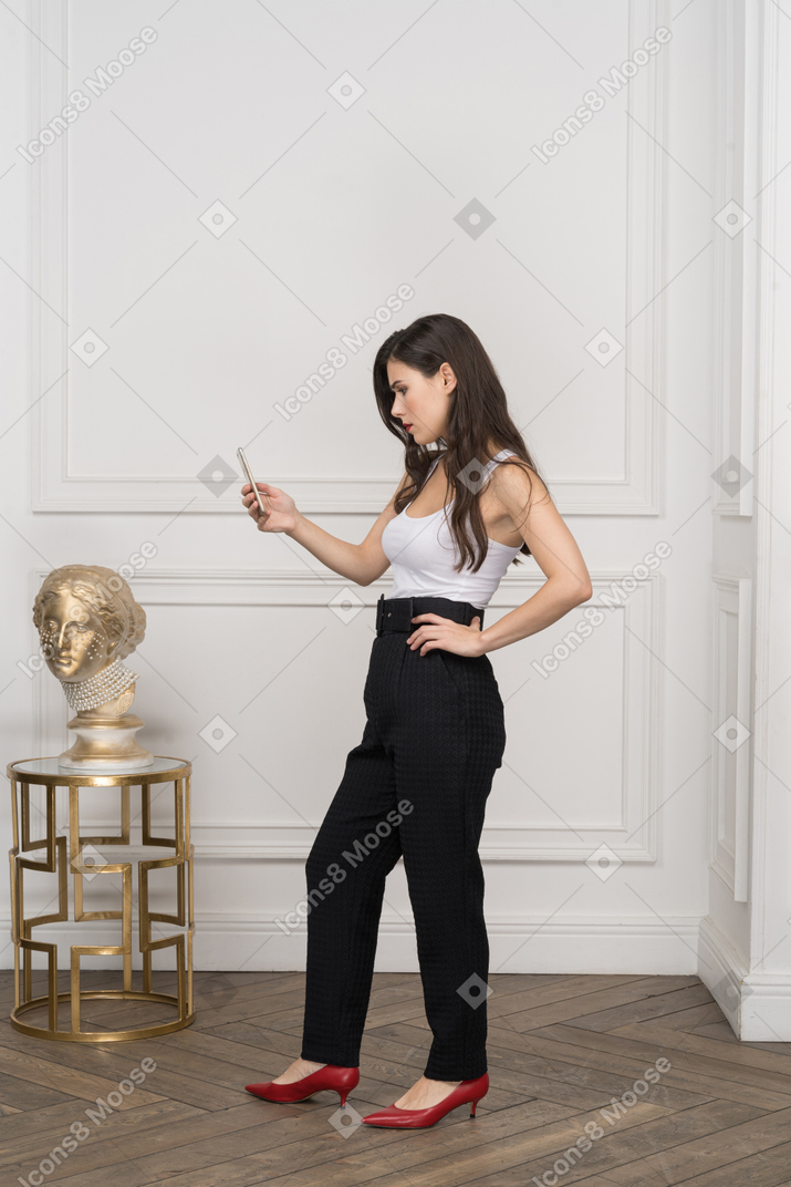 Side view of a young female looking at her phone and putting hand on hip near golden greek sculpture