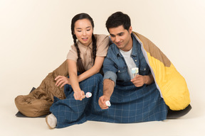Jovem casal interracial sentado no saco de dormir e segurando marshmallows
