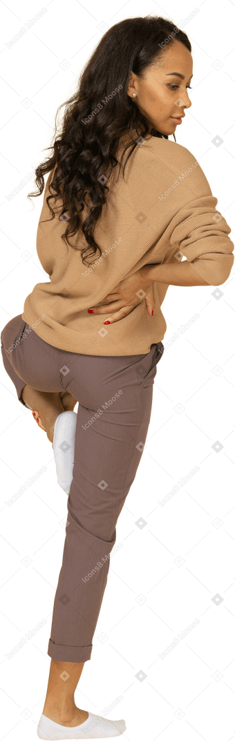 Вид сзади в три четверти темнокожей молодой женщины, которая кладет руку на бедро, поднимая ногу