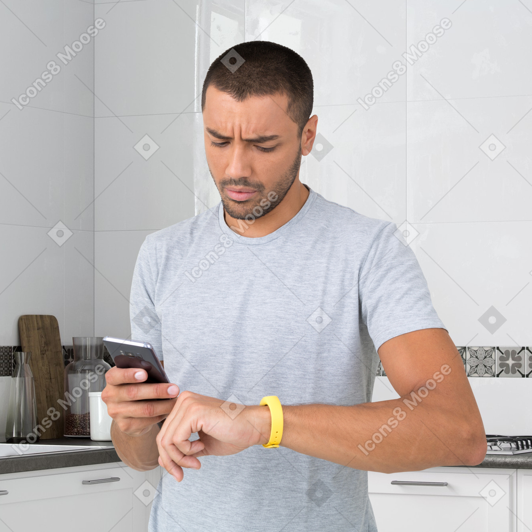 A man standing in a kitchen looking at his cell phone and a smartwatch