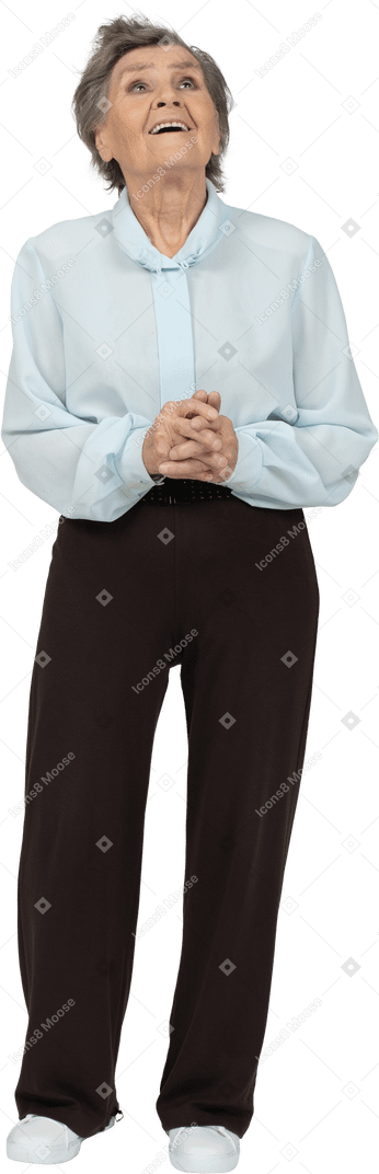 Front view of a happy old female in blouse and trousers holding hands together and looking up