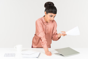 Mad looking young indian office worker standing near office desk and holding papers