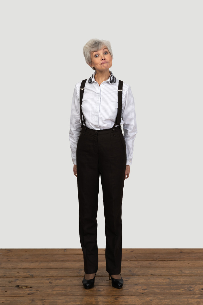 Front view of an old surprised female in office clothes grimacing with her hands behind back