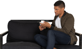 Three-quarter view of a young dreaming man sitting on a sofa with a cup of coffee
