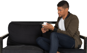 Dreiviertelansicht eines jungen träumenden mannes, der mit einer tasse kaffee auf einem sofa sitzt