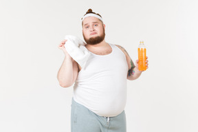 Разочарованный толстяк в спортивной одежде держит бутылку безалкогольного напитка и полотенце