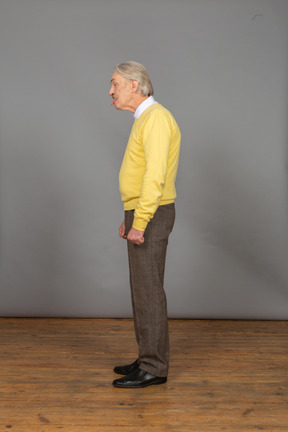 Vista lateral de un anciano curioso en jersey amarillo mostrando la lengua y mirando a un lado