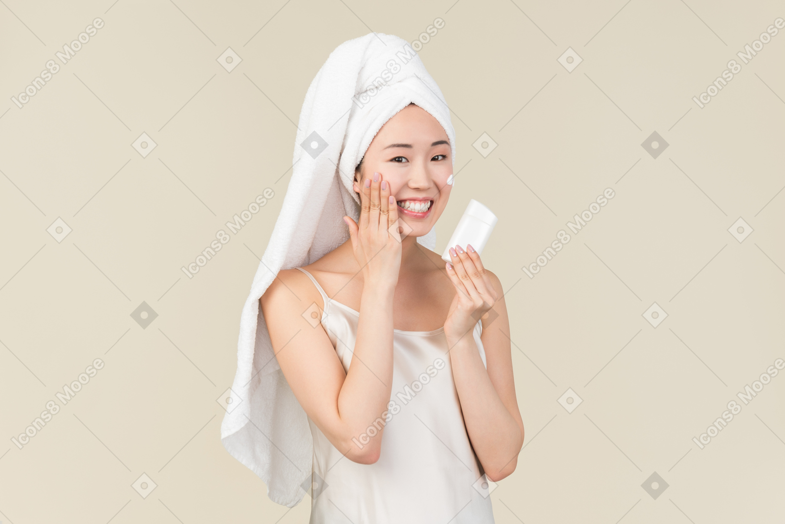 Улыбающаяся азиатская девушка с волосами, завернутыми в полотенце, наносит крем