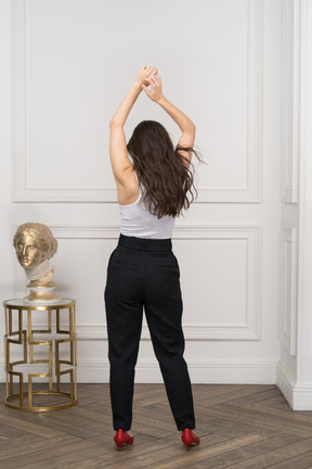 Vista de trás de uma jovem mulher levantando as mãos ao lado de uma escultura grega dourada