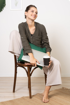 Вид спереди улыбающейся молодой женщины, сидящей на стуле и держащей свой ноутбук и чашку кофе