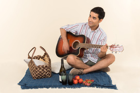 Junger kaukasischer kerl, der nahe picknickkorb auf der decke sitzt und gitarre spielt