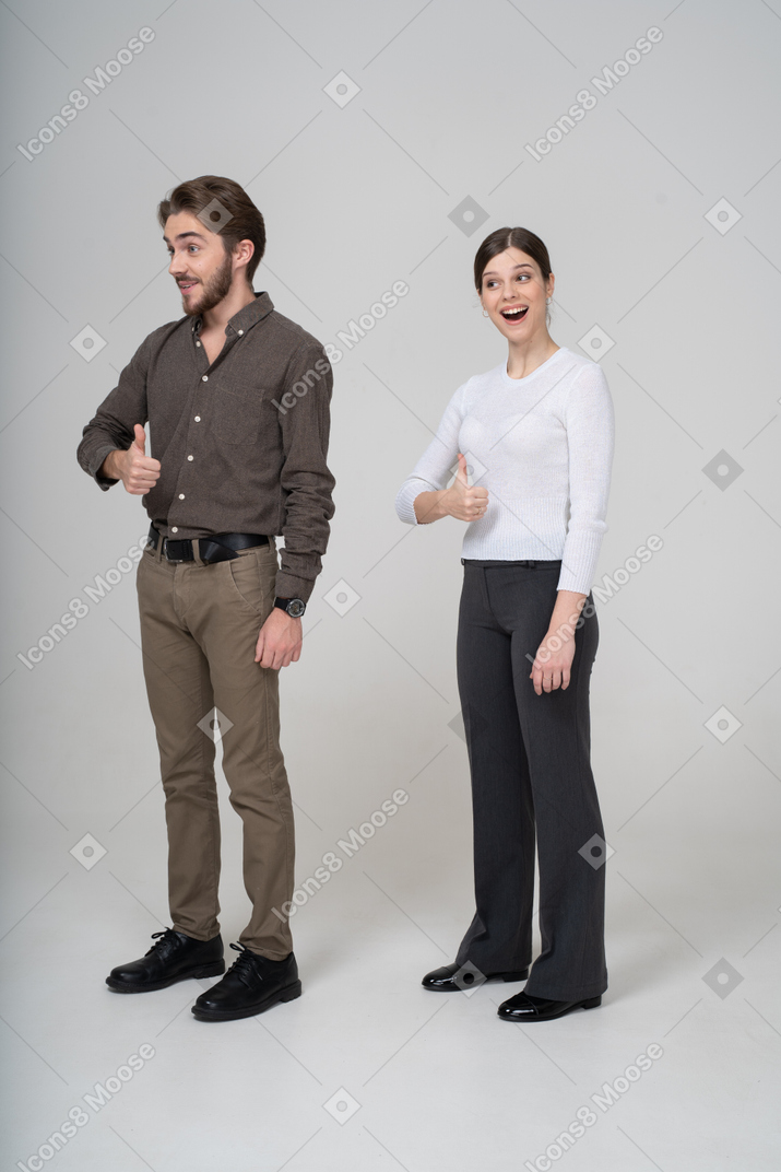 Vue de trois quarts d'un jeune couple joyeux en vêtements de bureau montrant le pouce vers le haut