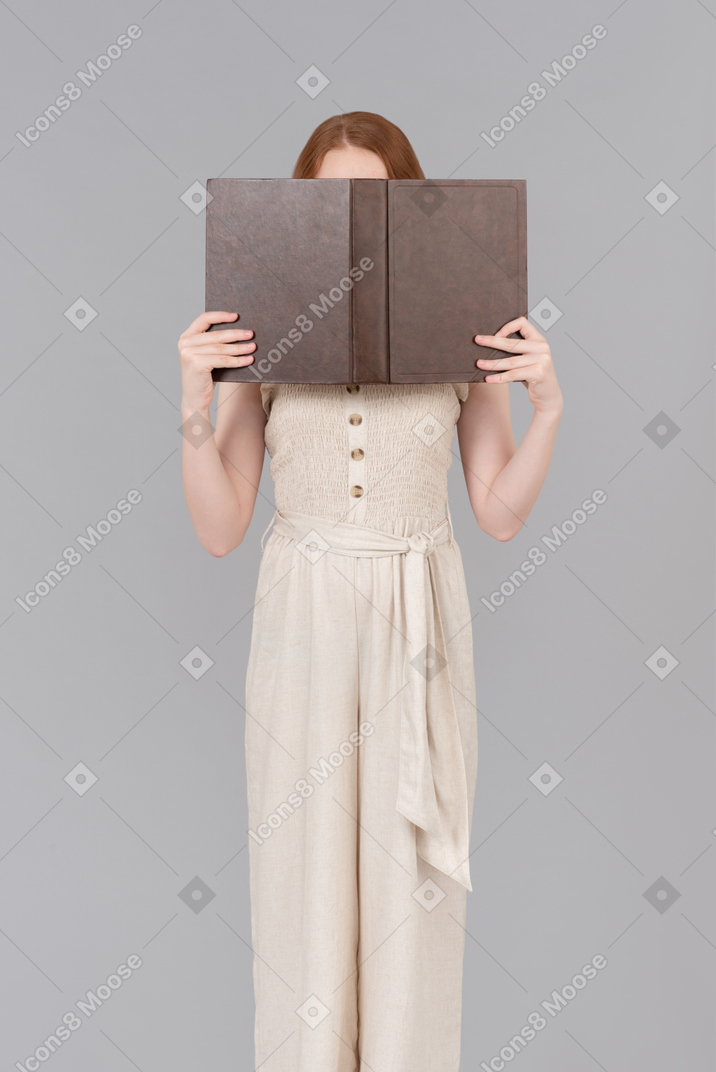Teenage girl dressed in beige overalls closing face with the book