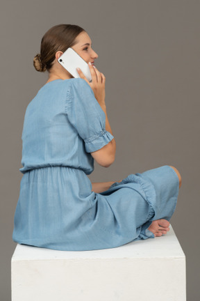 年轻女子坐在立方体上，用智能手机说话的侧背视图