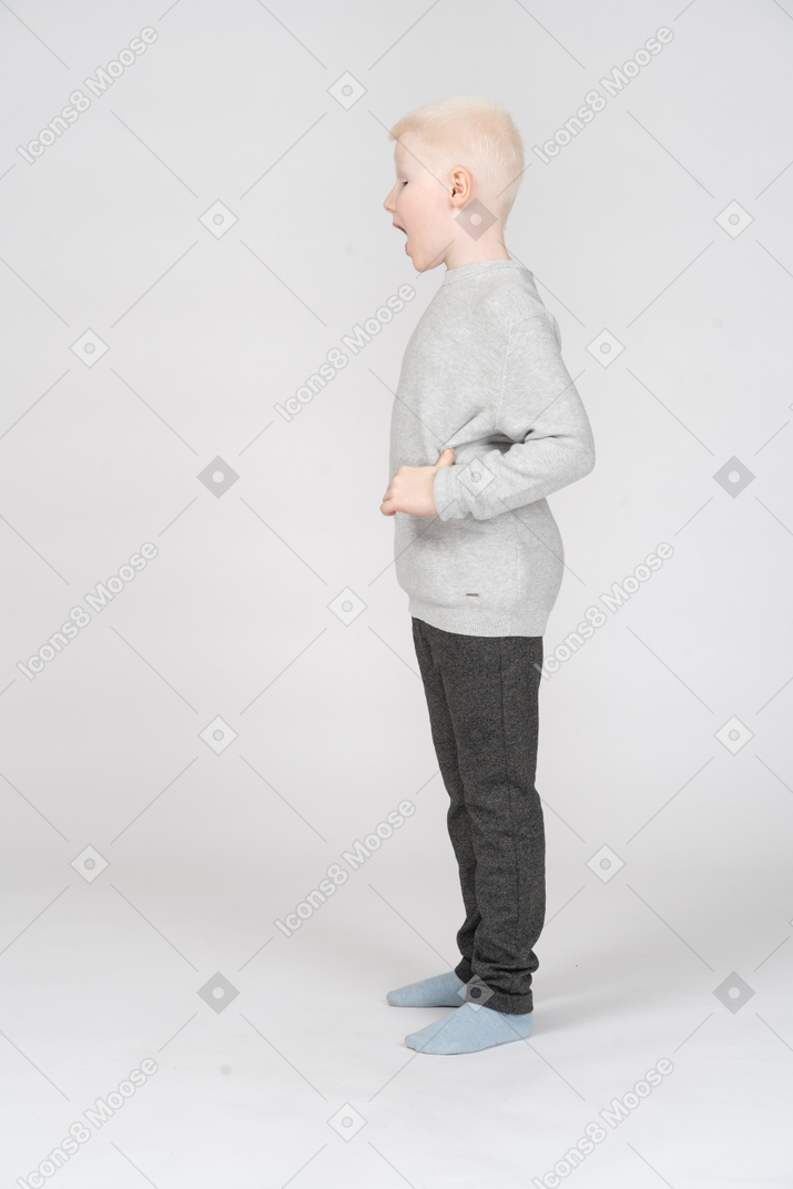 Side view of a kid boy in casual clothes opening mouth widely and putting hand on hip