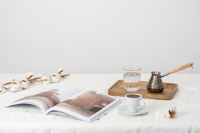 Cezve and glass of water on the wooden tray, cup of coffee and magazine and cotton branch