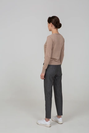Three-quarter back view of a young lady standing still in pullover and pants