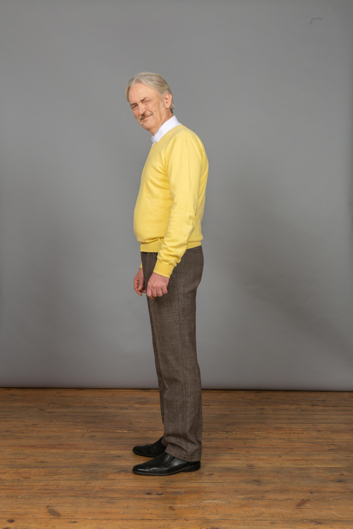 Side view of an old displeased man in yellow pullover narrowing eyes and looking at camera