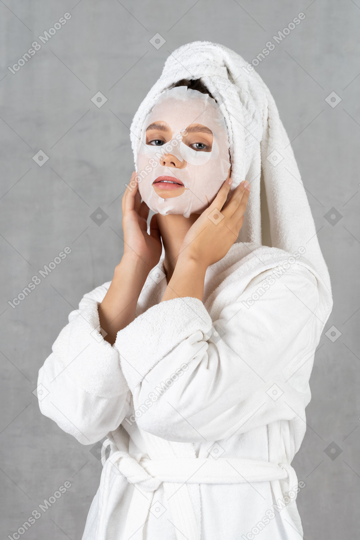 Woman in bathrobe with face mask on touching her face