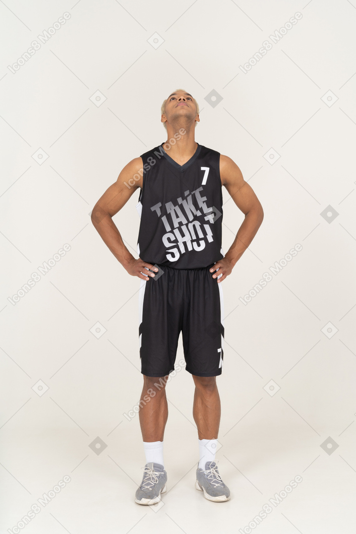 Vista frontal de um jovem jogador de basquete colocando as mãos nos quadris e olhando para cima