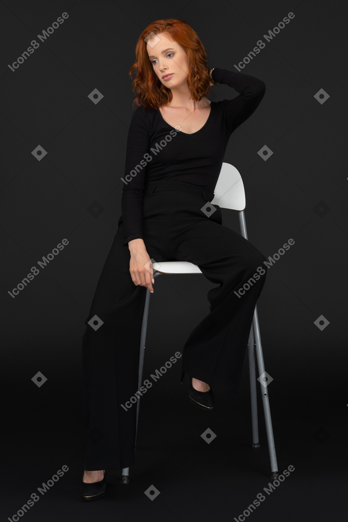 A frontal view of the sexy young woman sitting on the tall grey chair and adjusting her hair