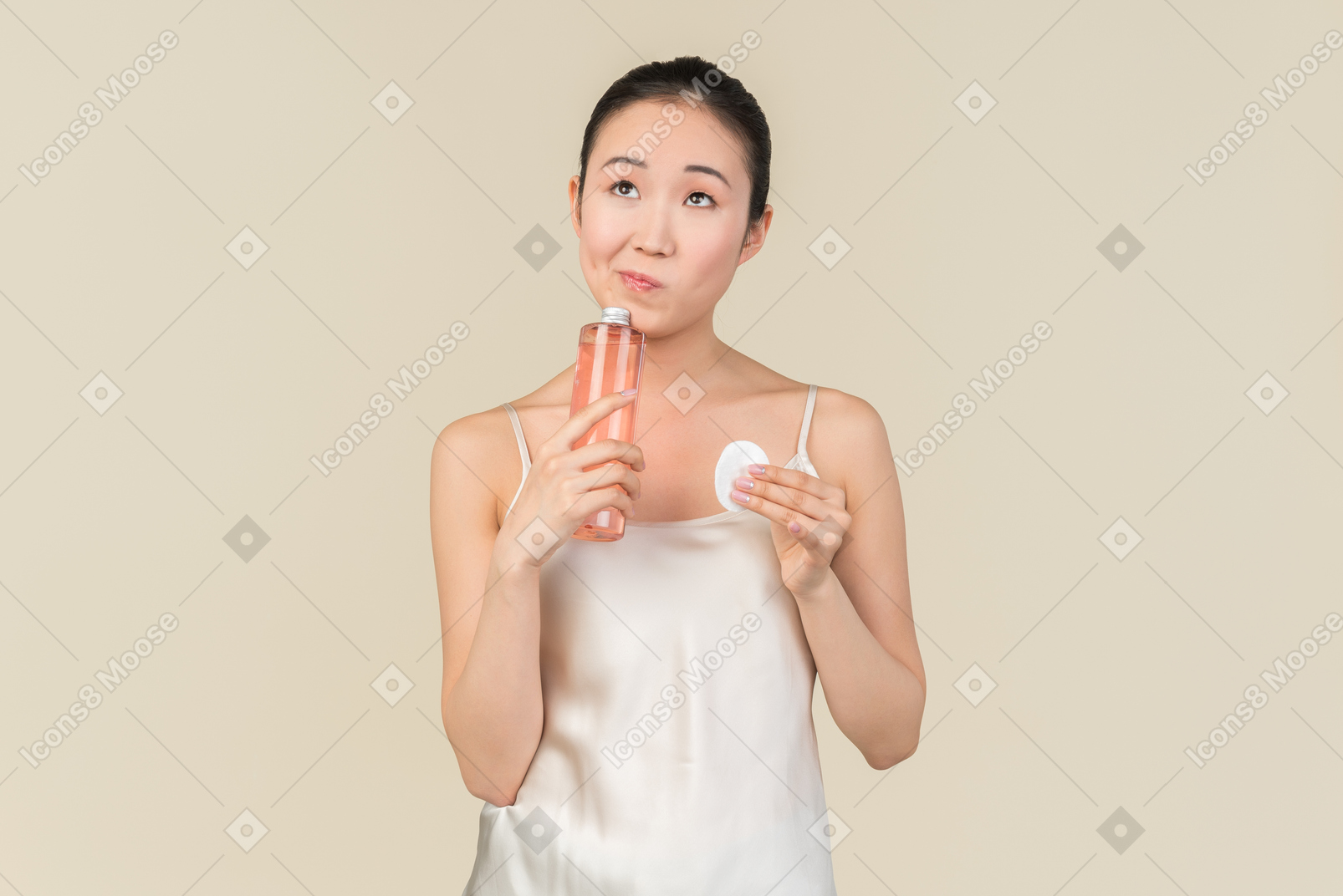 Pensive young asian woman holding cosmetic bottle and cotton pad